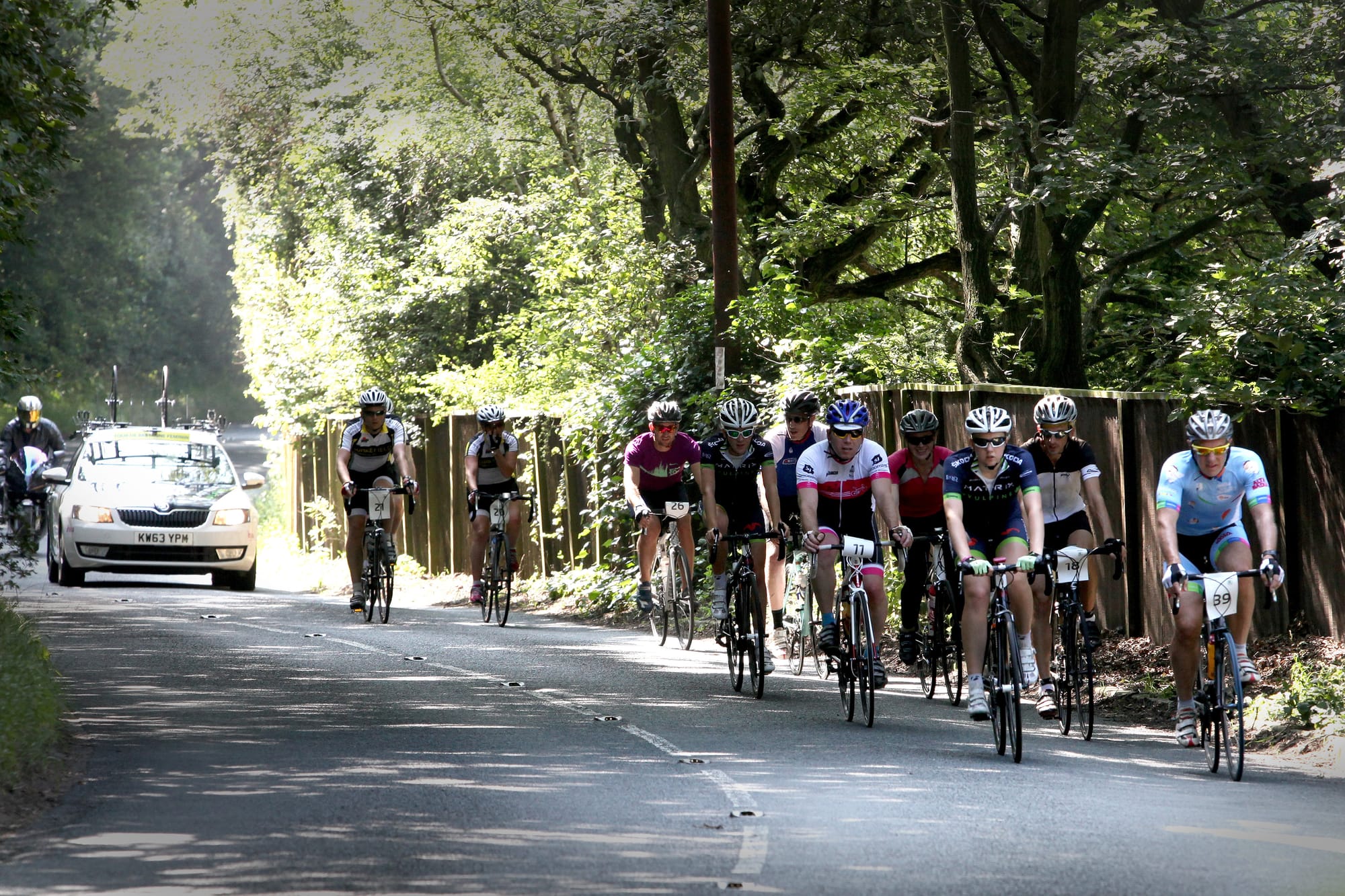 A group of cyclists on a winding, leafy country road, followed by a support car