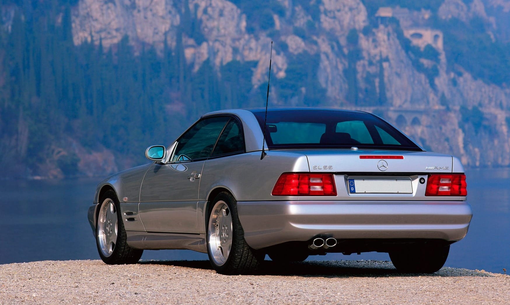 An R129 generation of Mercedes SL photographed from the rear three-quarter angle, and parked next to a lake with mountains in the background 